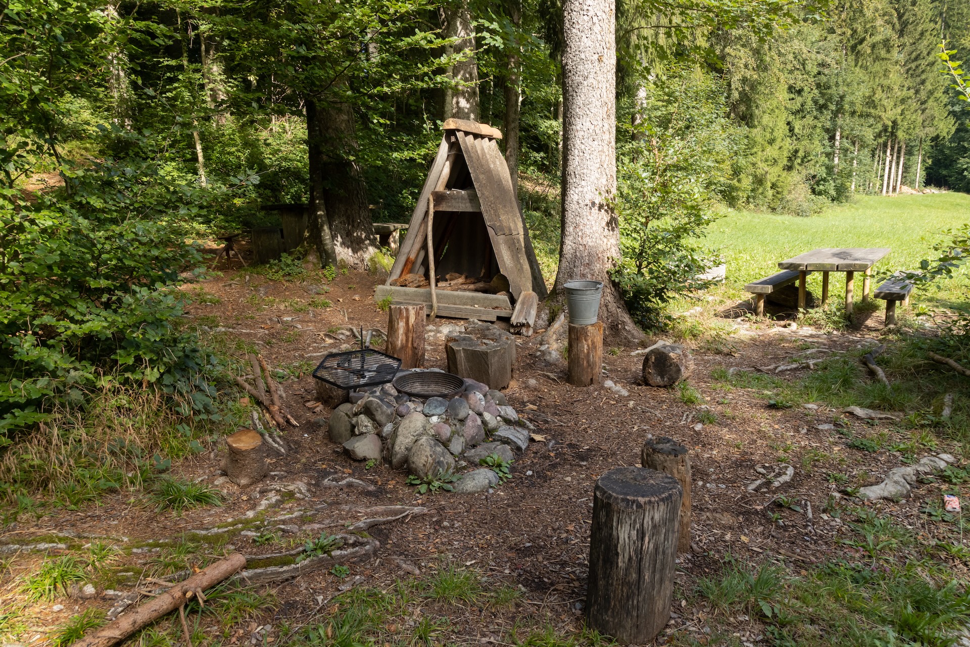 Stein-Feuerstelle mit Grill im Wald mit überdachten Holzscheiten daneben und einem Tisch mit Bänken